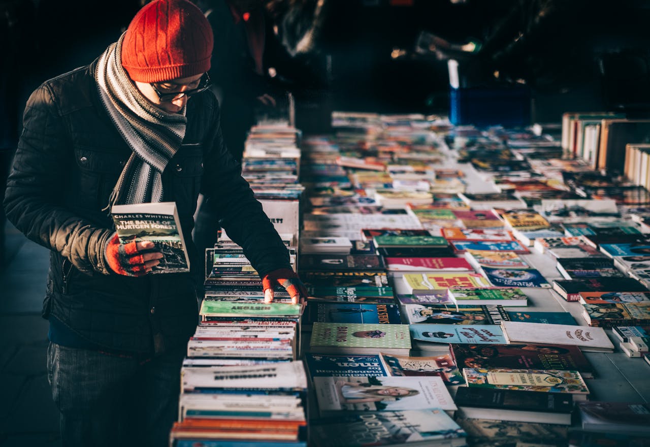 A man dressed warmly in a beanie and scarf browses through books at an outdoor evening market.