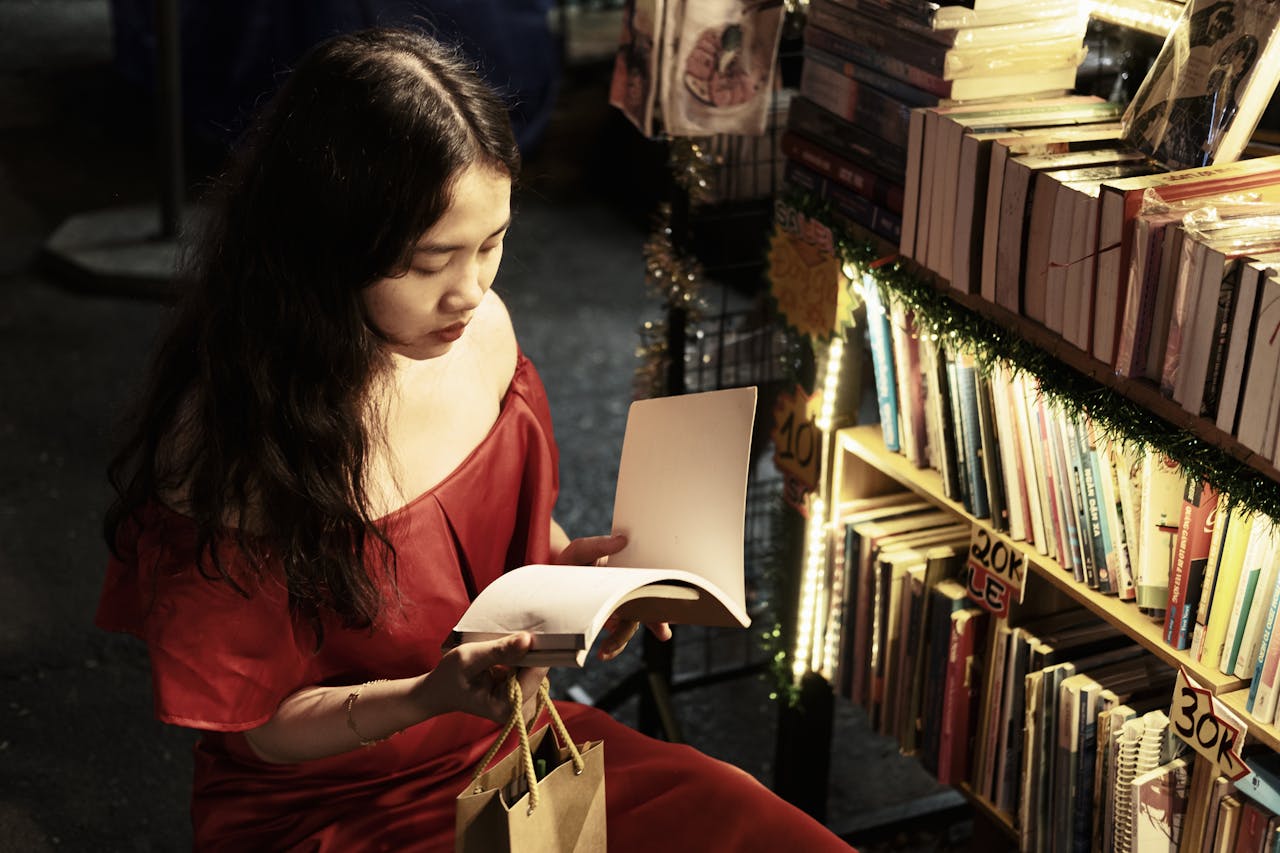 Woman in red dress reading a book at a dimly lit night market bookstall, surrounded by books.
